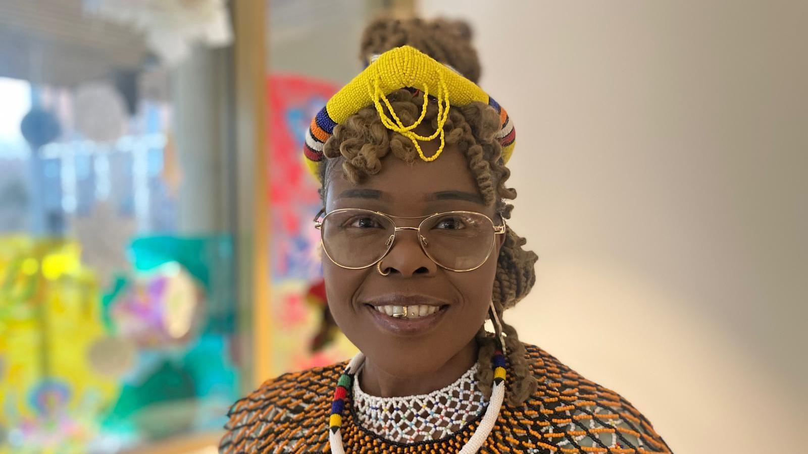 A woman with gold glasses and traditional South African attire smiles at the camera. Behind her, a blue and yellow window display is blurred.