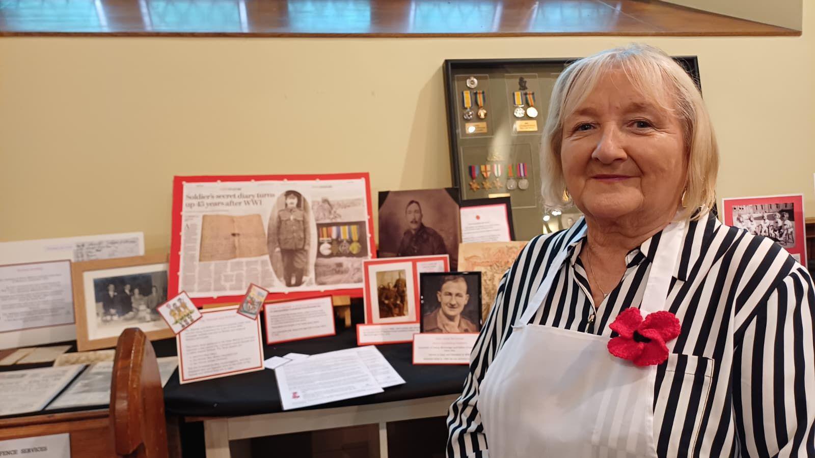 Anne is wearing an apron with a poppy on it. Her top is black and white stripes. Behind her is war memorabilia.