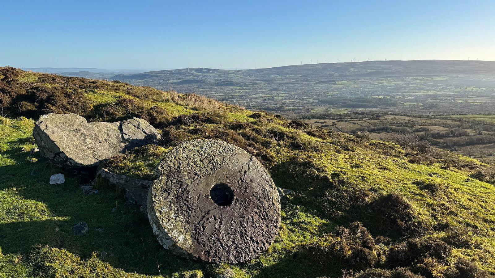 An old millstone sits on a hill in Kinawley, County Fermanagh, Northern Ireland. The scenery is stunning - a blue sky and green fields are seen for miles.