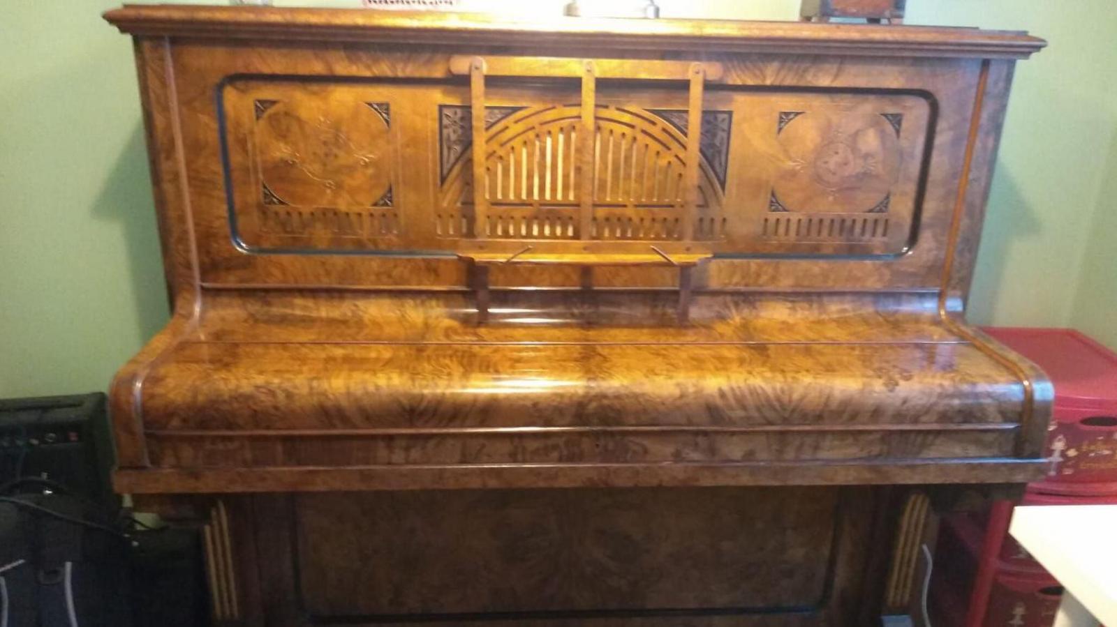 An upright piano made of light patterned wood, standing against a sage green wall.