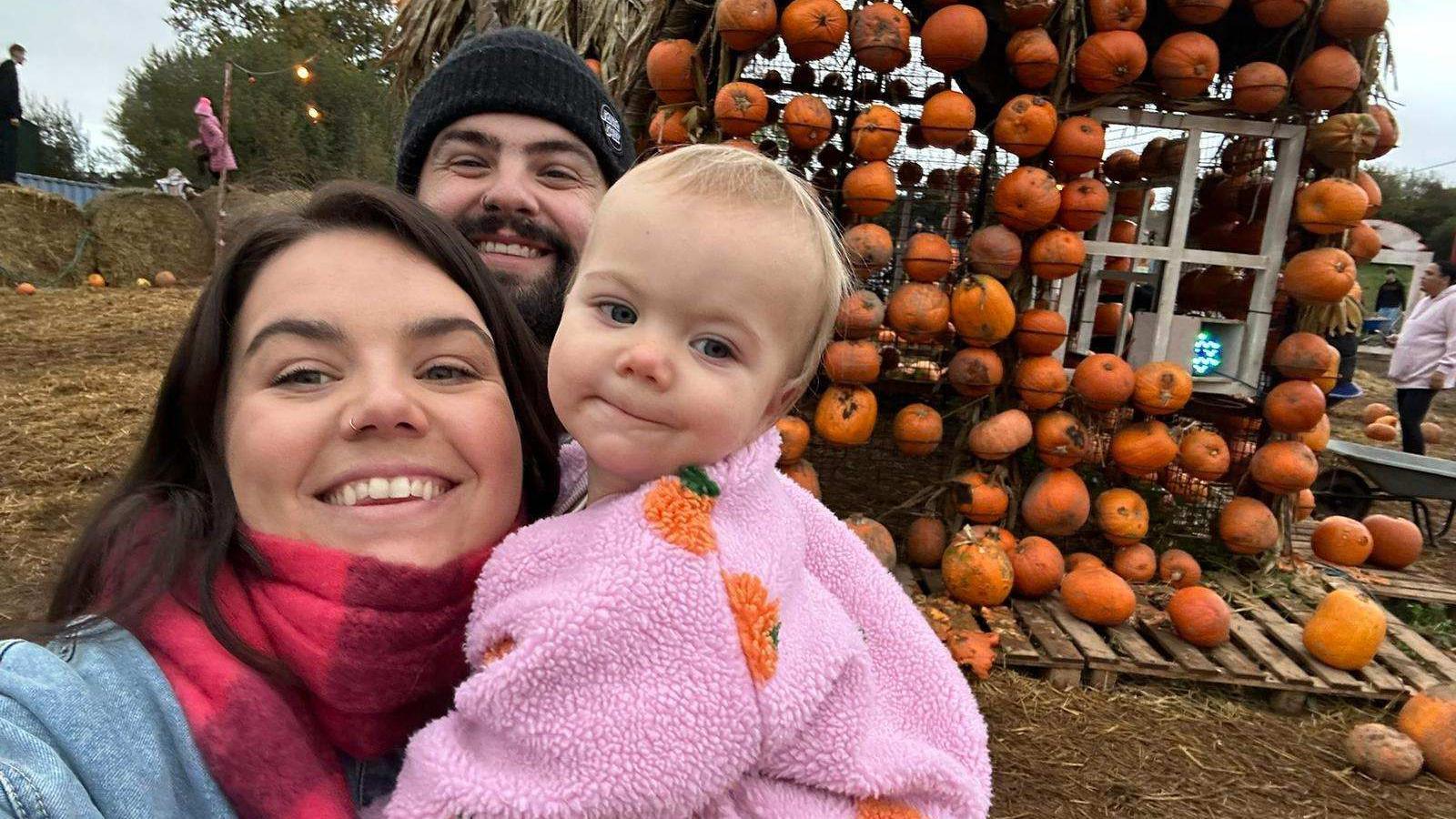 A mum holds her toddler, who is wearing a pink fleece with pumpkins on it. A man in a woolly hat - her father - can be seen behind the pair. There is a display on pumpkins in the background in a muddy field. 