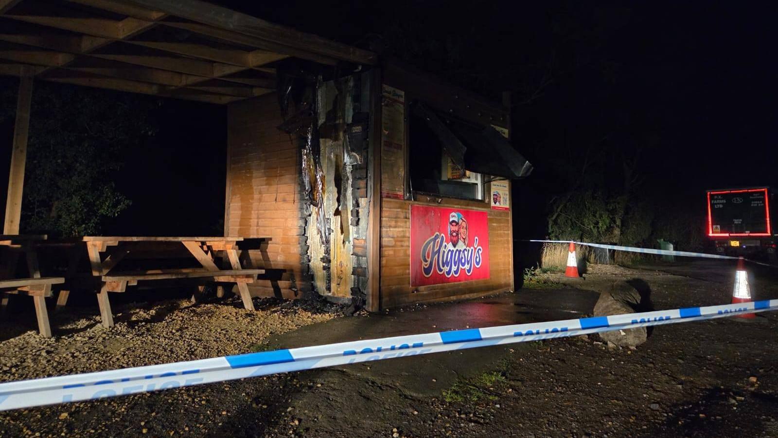 A blue and white police cordon around a brown shed- damaged by fire.