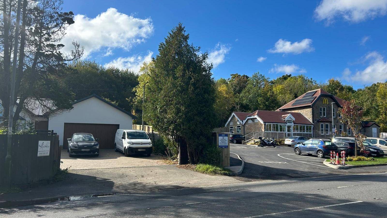 Two cars are parked in front of a garage to the left of a fence while on the other side is a car park and McDonald's restaurant