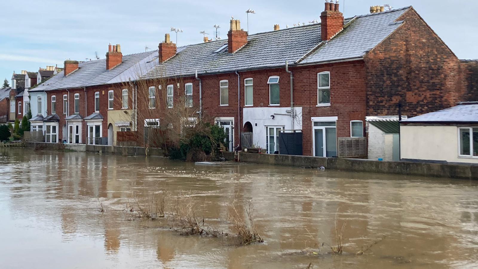 A residential street will with brown flood water in front of a row of terraced red-brick houses.