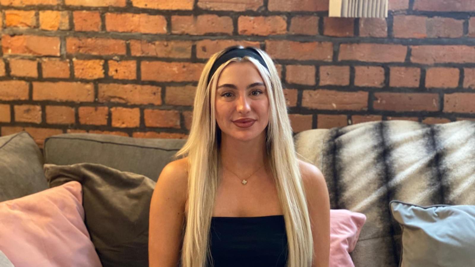 A young woman with long straight blonde hair held back by a thick black headband sits on a grey sofa next to a bare brick wall. She's smiling slightly.