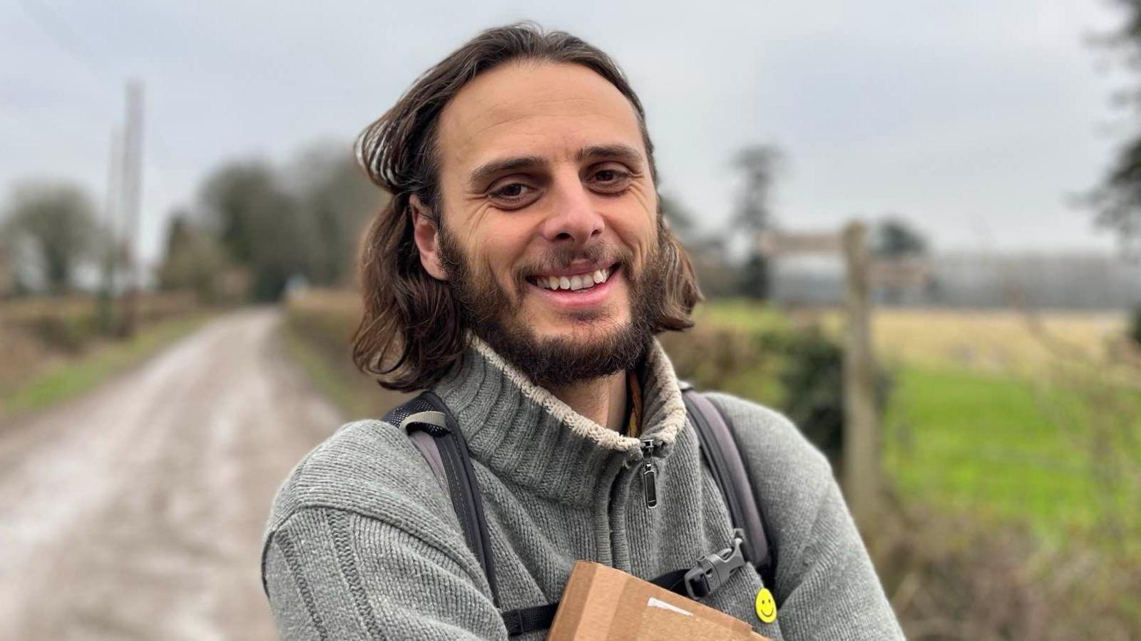 A man with shoulder-length brown hair and beard looking and smiling at the camera. He is wearing a grey turtle neck jumper and is standing in front of a country road.