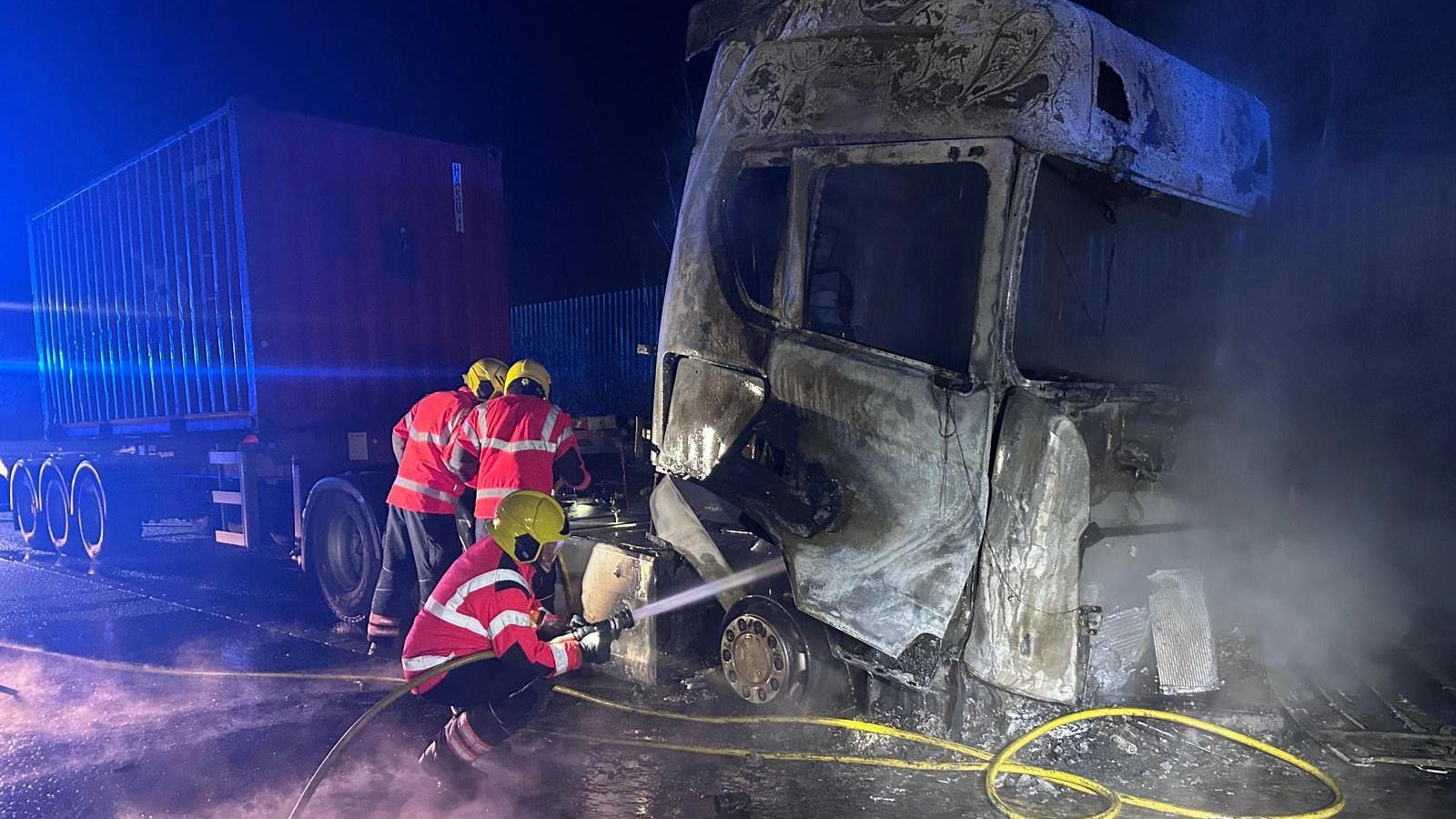 Firefighters extinguishing a lorry which has been burnt due to a fire. They wear bright firefighting uniform and use a water hose to douse the vehicle. 