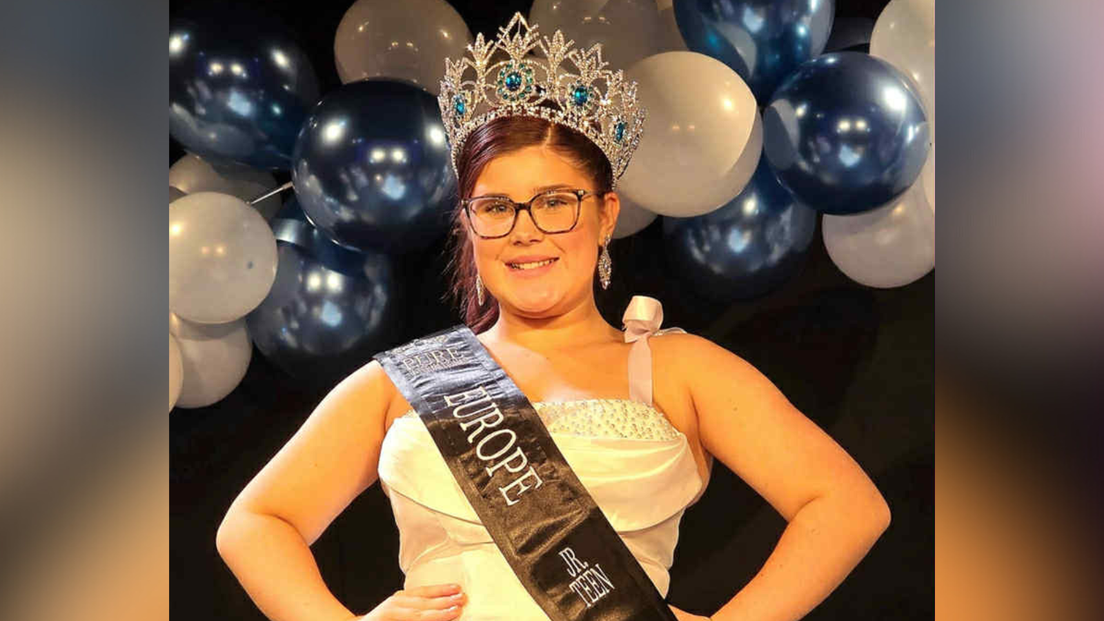 Bella stands on stage wearing crown and a sash and a white dress. She has brown hair tied up and is wearing glasses and silver earrings. A balloon arch is visible in the background.