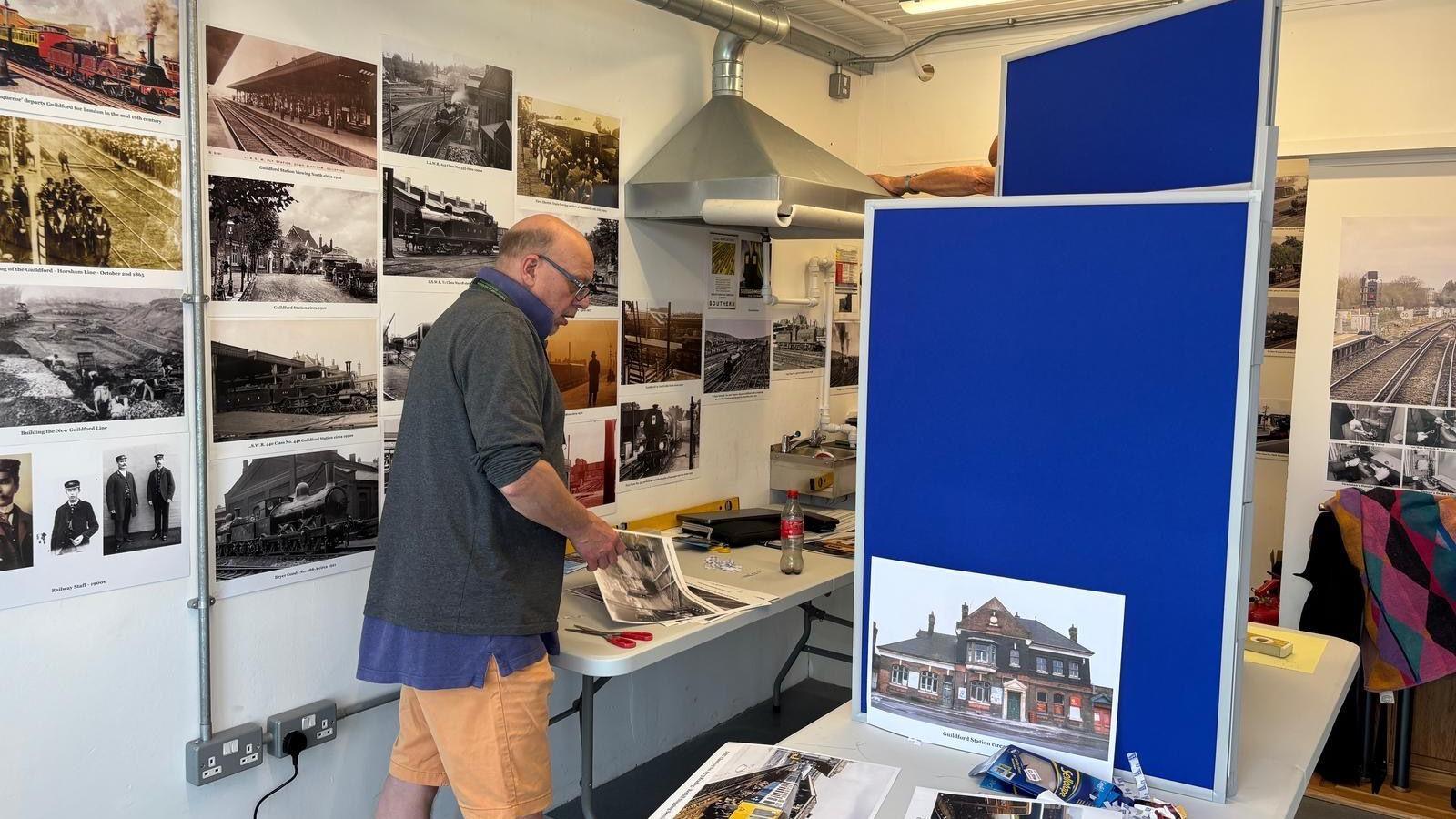 A man is picking up a printed archive image to display in an exhibition. Images of railwaymen in uniform, train tracks and Guildford station are placed on the walls of the white room. They range from historical black, white and sepia-filtered photographs to modern ones in full colour.
