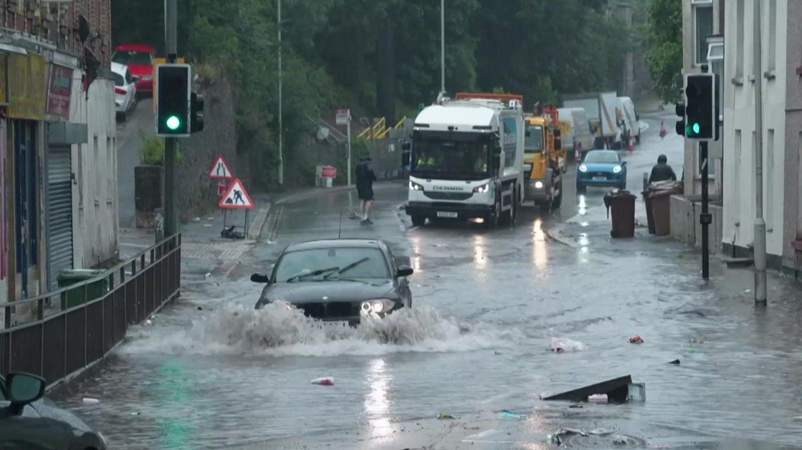A car driving through flood water, half submerged. There is some rubbish floating in th water.