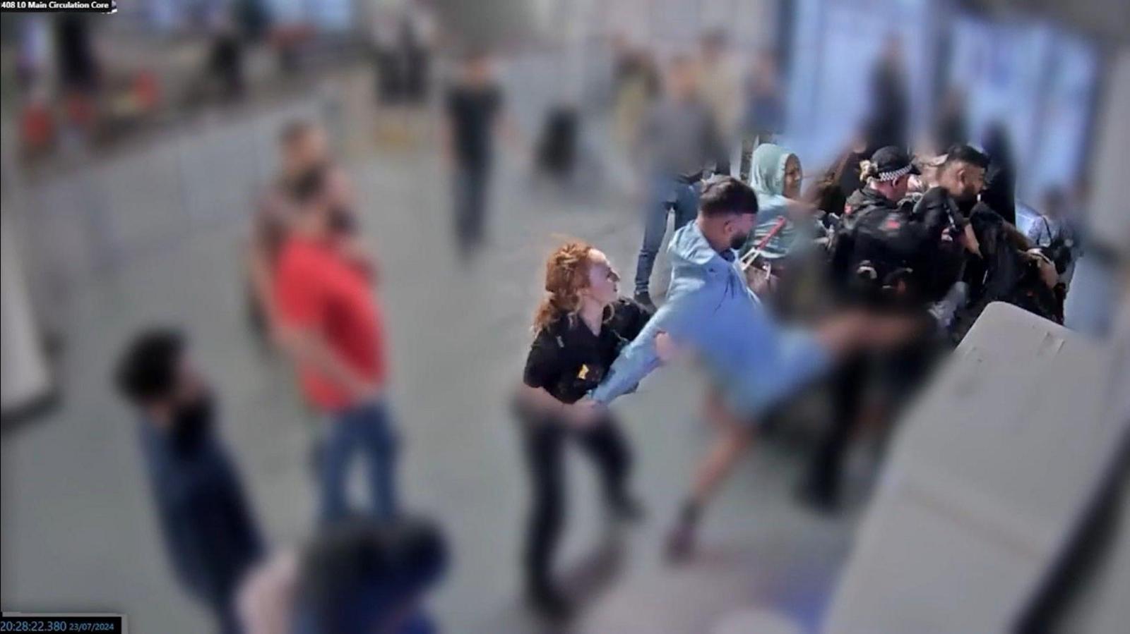 A man in blue kicks in the direction of a police officers while a police officer with red hair holds his arms behind him at the car park pay stations at Manchester Airport.
