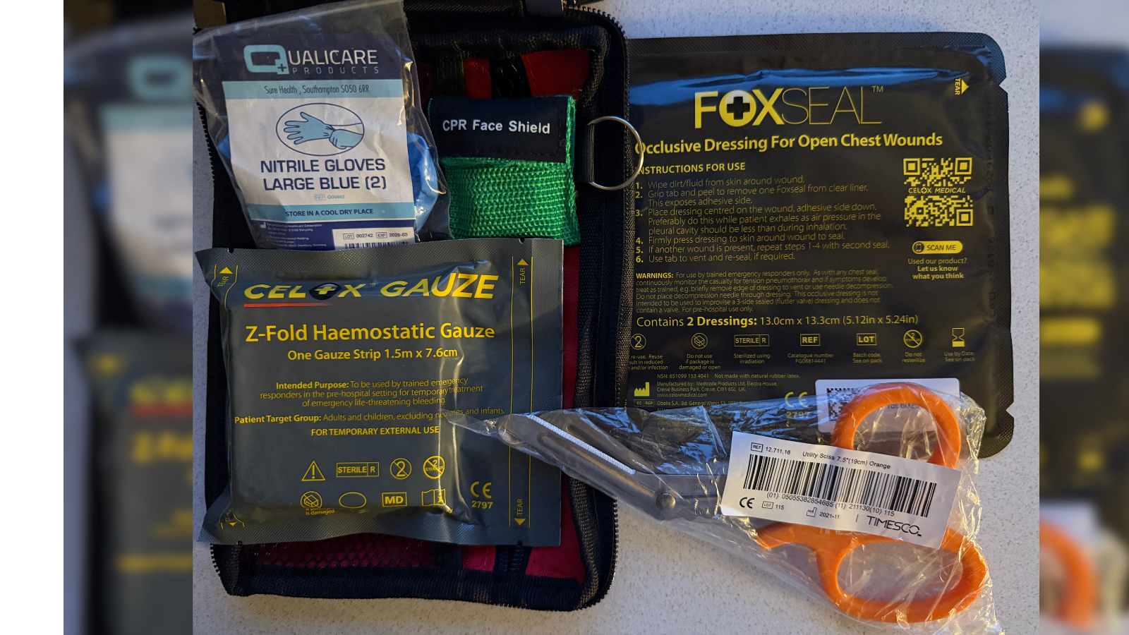 Emergency equipment, including scissors, haemostatic gauze, gloves, a face shield and a seal for open chest wounds, are shown inside an open bleed kit.