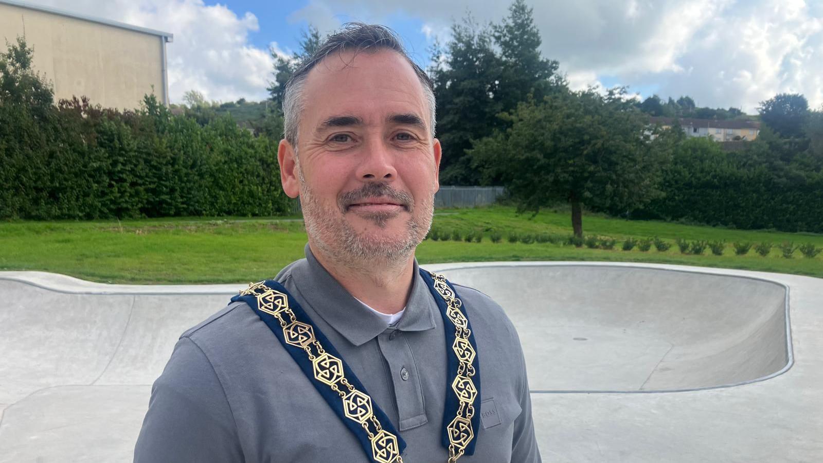 This photo shows a man standing outdoors at a skatepark, wearing a grey collared shirt and a ceremonial gold and blue chain. Trees, bushes, and grassy areas are visible in the background under a partly cloudy sky.