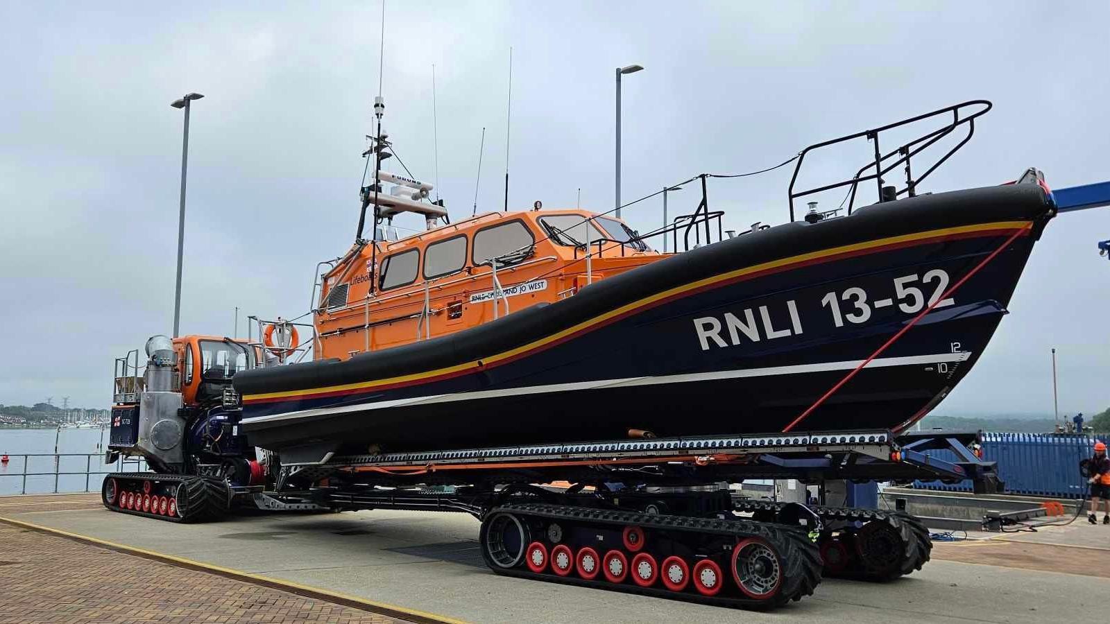 A lifeboat, which has an orange cabin and a navy hull with the lettering "RNLI 13-52" on it. The boat is on a platform made of wheels at a dockyard, with the sea behind it. It is a grey, cloudy day.