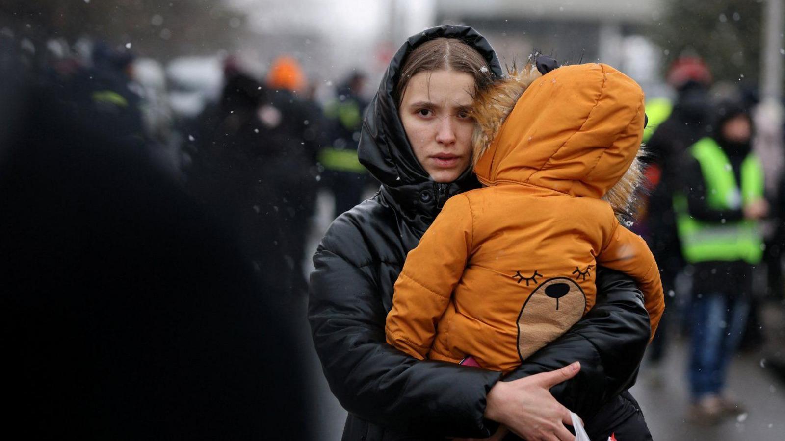 A woman in a black raincoat carrying a child at a border crossing near Ukraine. It is snowing. 