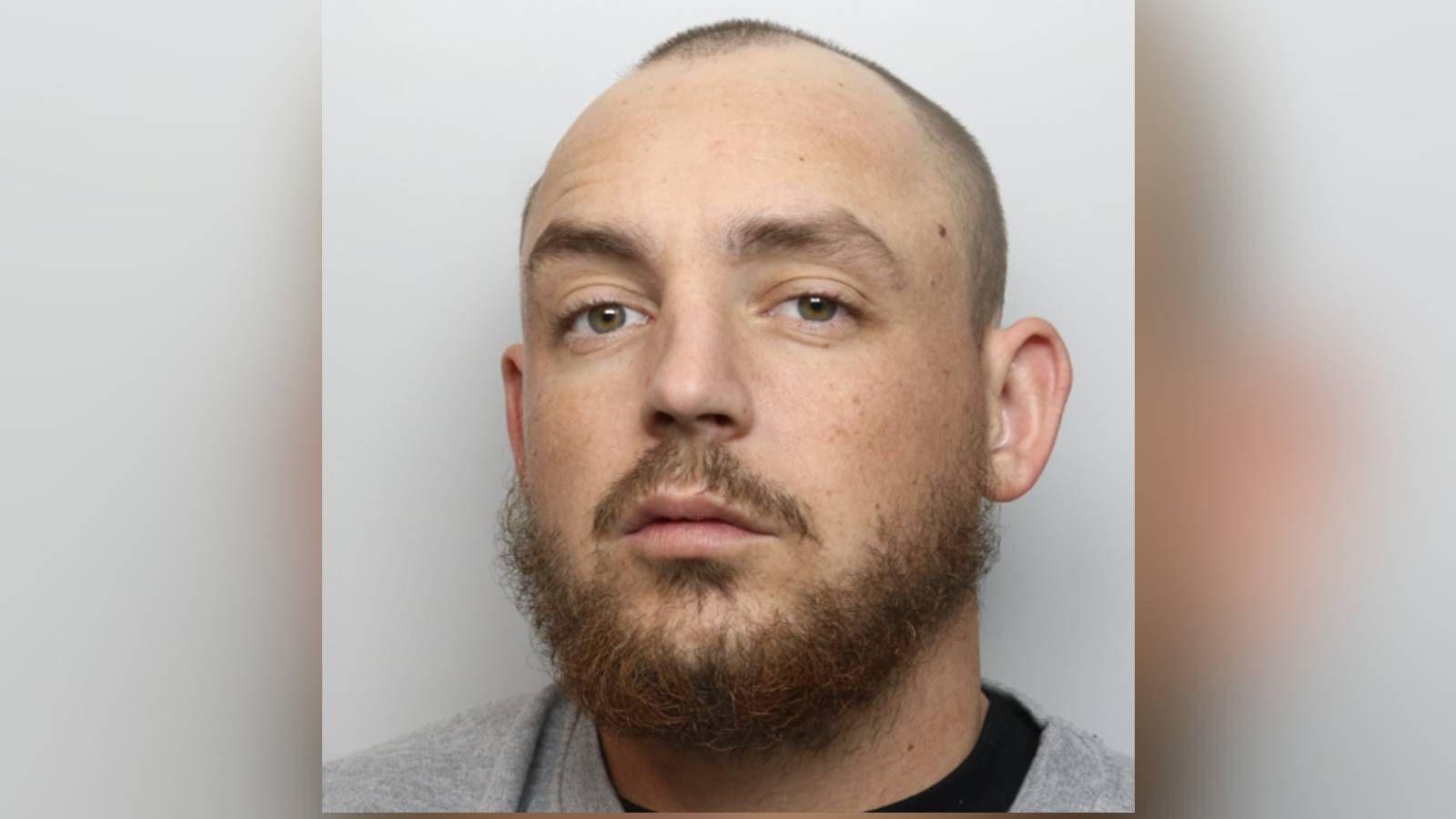 A police mugshot of a man with a shaven head and a brown beard and moustache, and wearing a grey top, with a black T-shirt underneath.