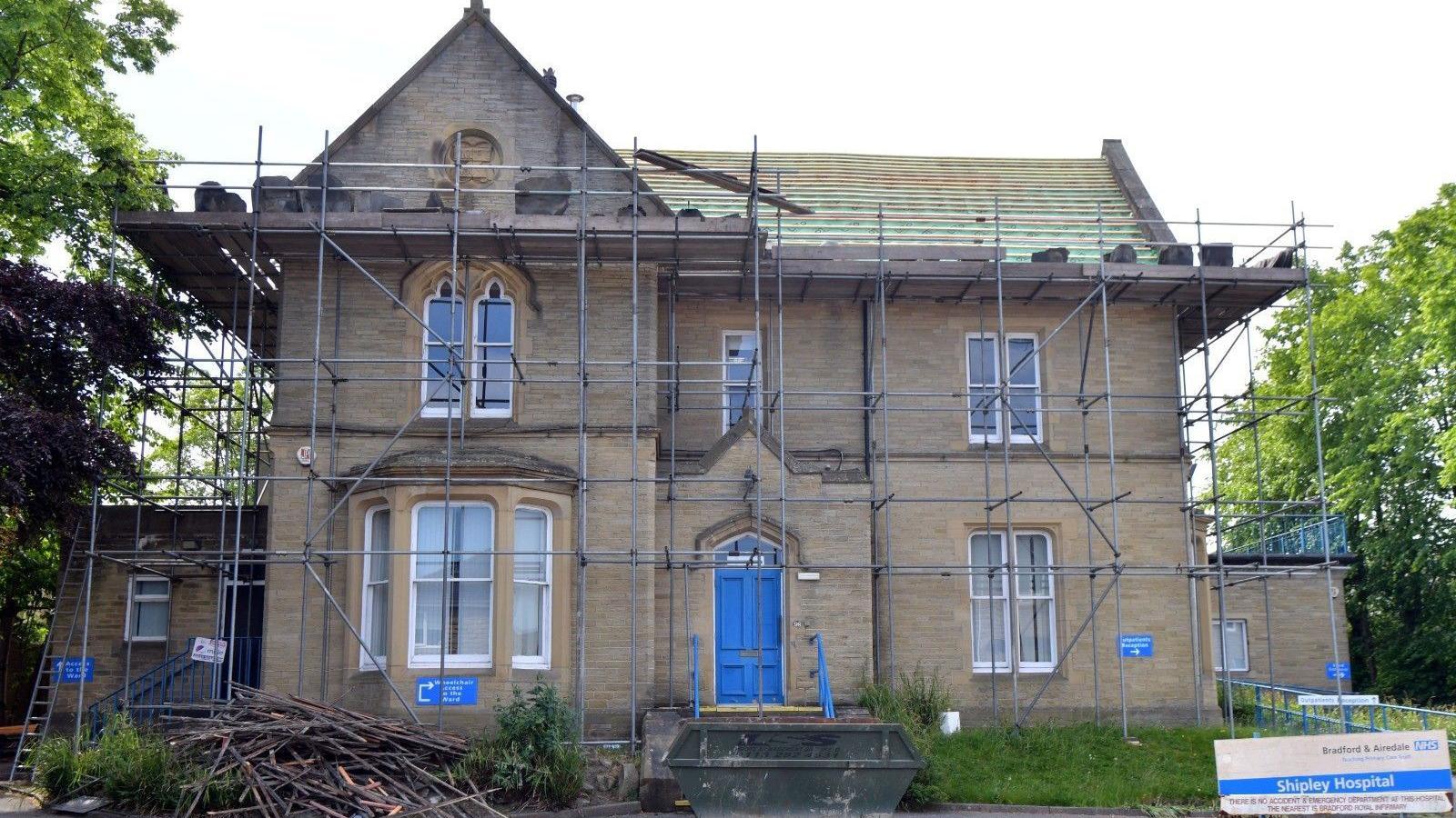 A sandstone Georgian hospital building with a bay window to the left of a bright blue front door. The pitched roof has been stripped of tiles and and there is scaffolding around the building, with a green skip at the front and a pile of discarded wood beside it. There is a old sign that says Shipley Hospital.