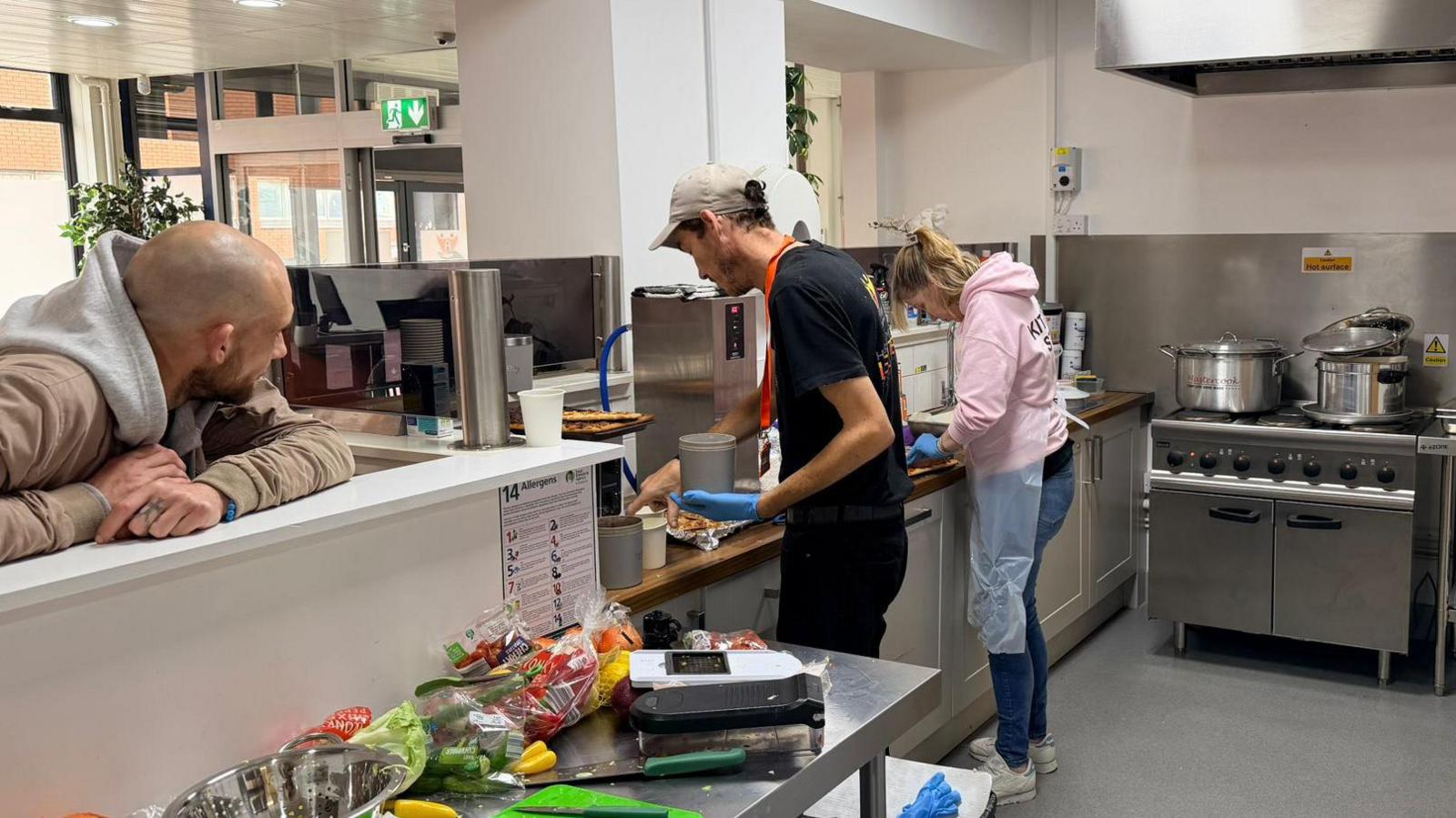 Two people working in a kitchen with a stove in the background and a man leaning on the counter