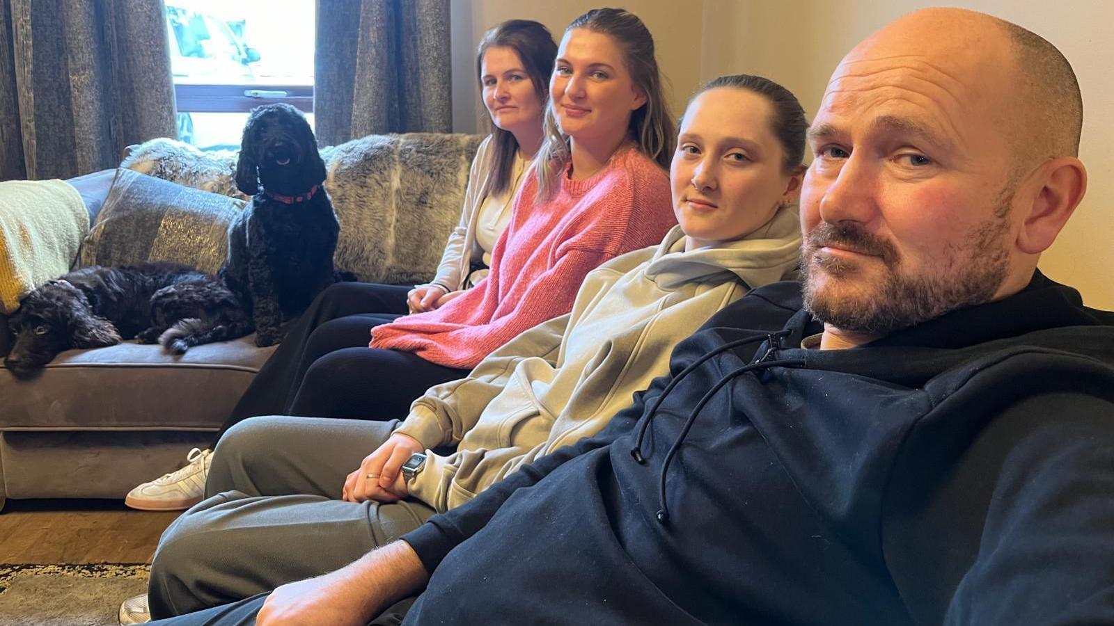 Lachlan is sitting on a sofa with his two daughters and his wife Leeann. The family's dogs have couch all to themselves.