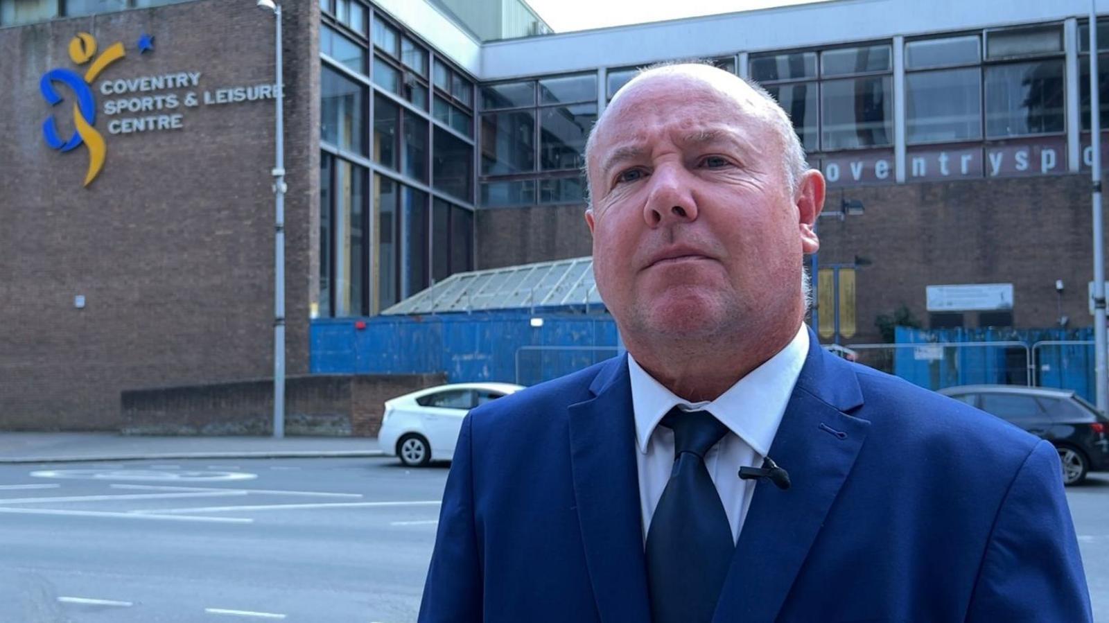 Councillor Jim O'Boyle stands in front of the closed Coventry Sports and Leisure Centre. He has short grey hair and is wearing a blue suit and tie and a white shirt.