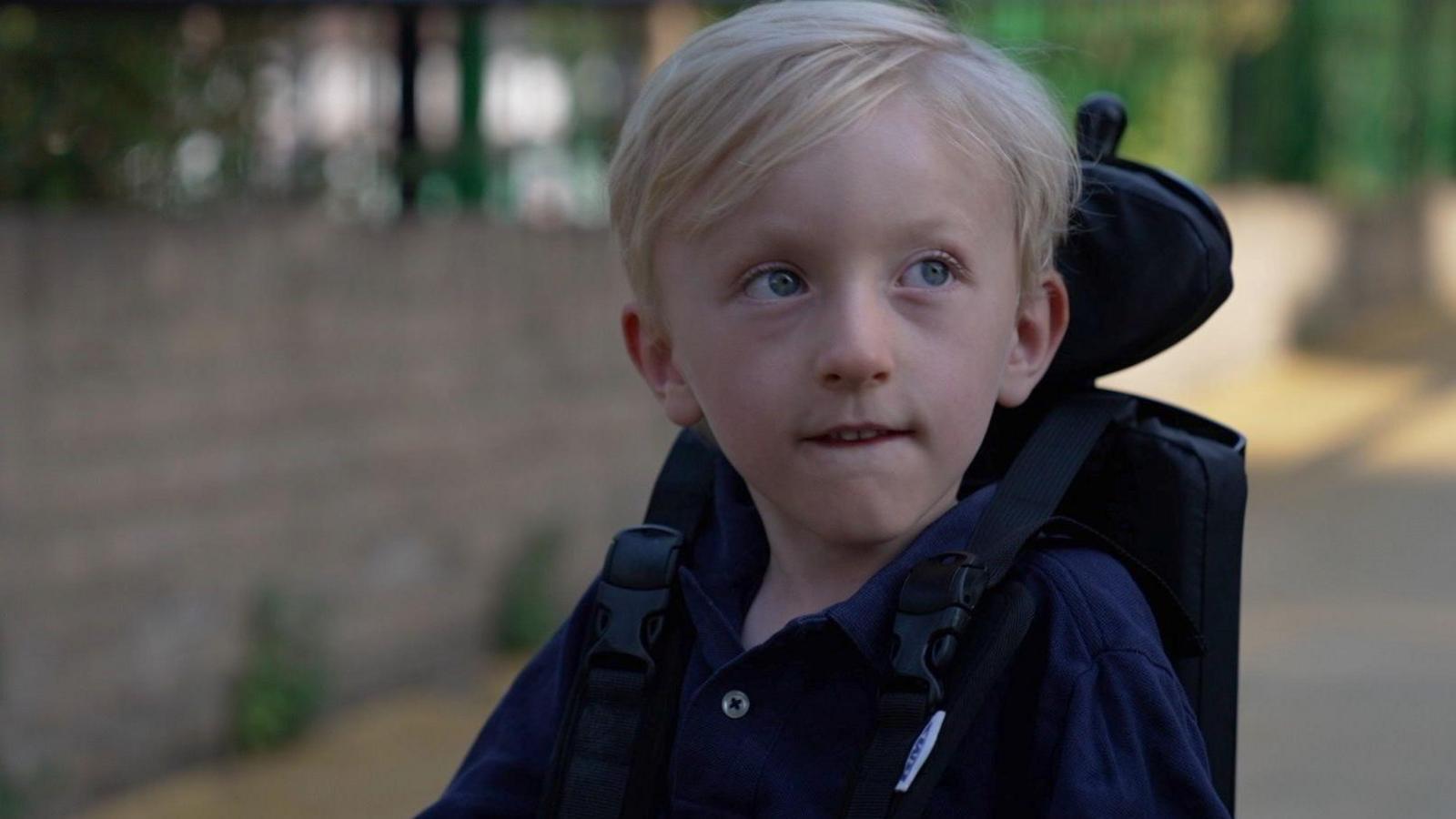 A young boy with blond hair and light coloured eyes, wearing a dark blue top and sitting in a wheelchair.