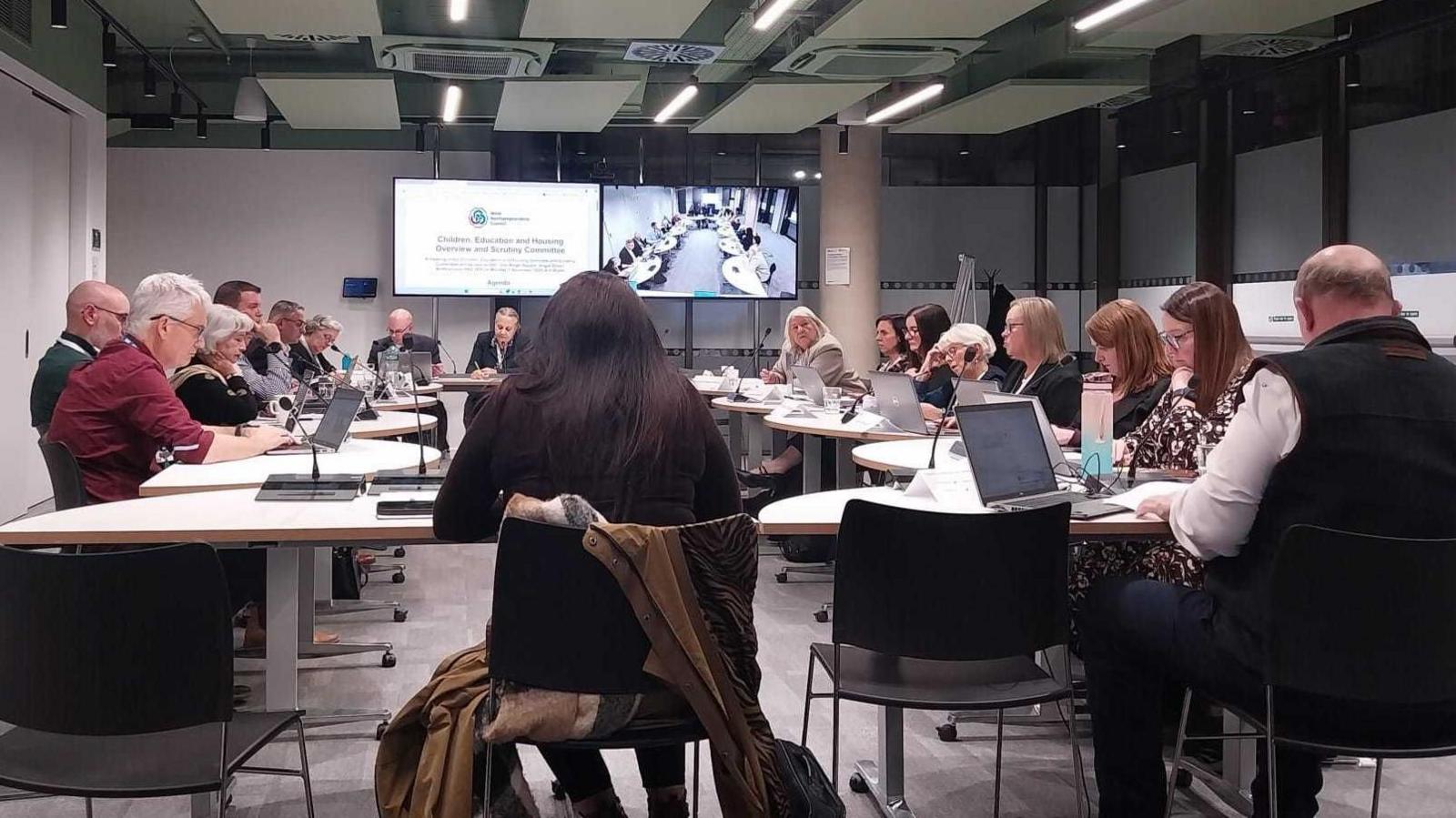 Councillors sitting around a table during a meeting. Many of them have laptops and microphones in front of them. Meeting papers are being shown on a large TV screen.
