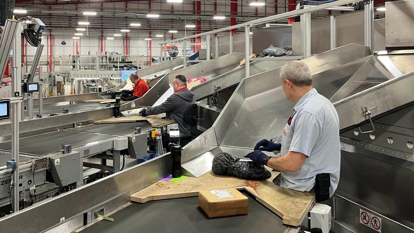 Three Royal Mail workers can be seen working at individual stations, preparing parcels for delivery before Christmas. The further man forward is wearing a red shirt, with the man in the middle wearing a grey jumper. The most visible man is wearing a short blue Royal Mail shirt and is preparing a black parcel for delivery. All three men are wearing gloves.