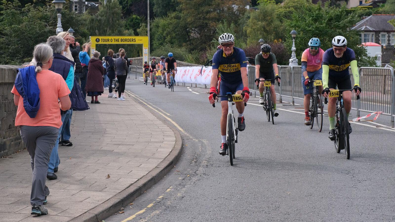 A number of cyclists crossing the finish line in Peebles on the Tour O The Borders - some spectators are lining the route