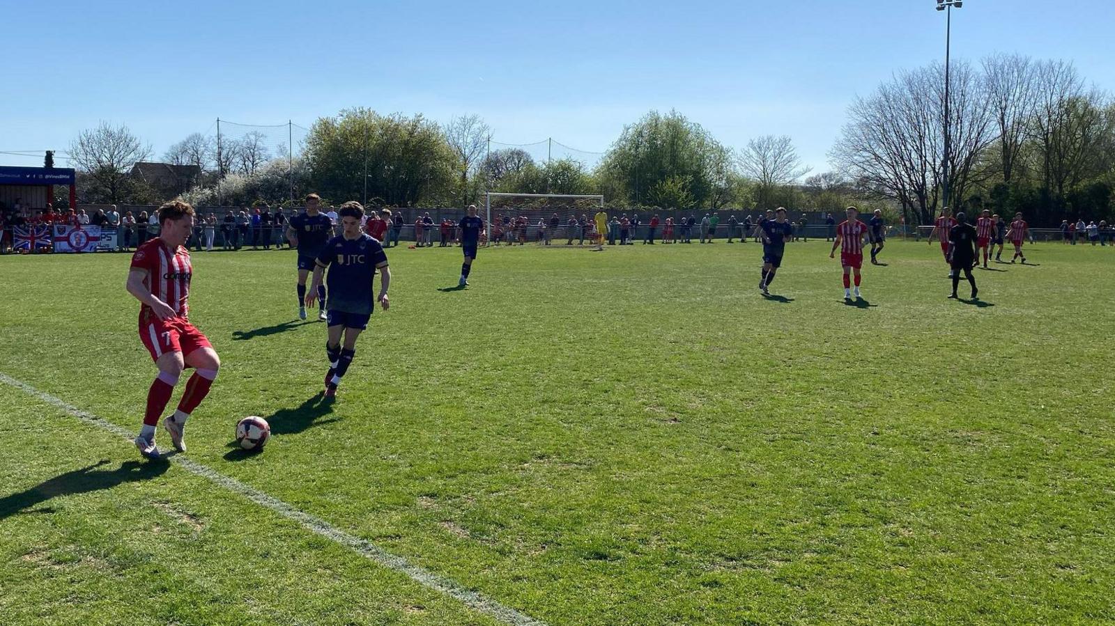A footballer wearing a red and white striped kit goes to kick a red and white ball with his left foot near the touchline along the side of a pitch. A player wearing a black kit is running towards him. Dozens of spectators are stood around the edge of the ground. Several other players and a referee are on the field of play.