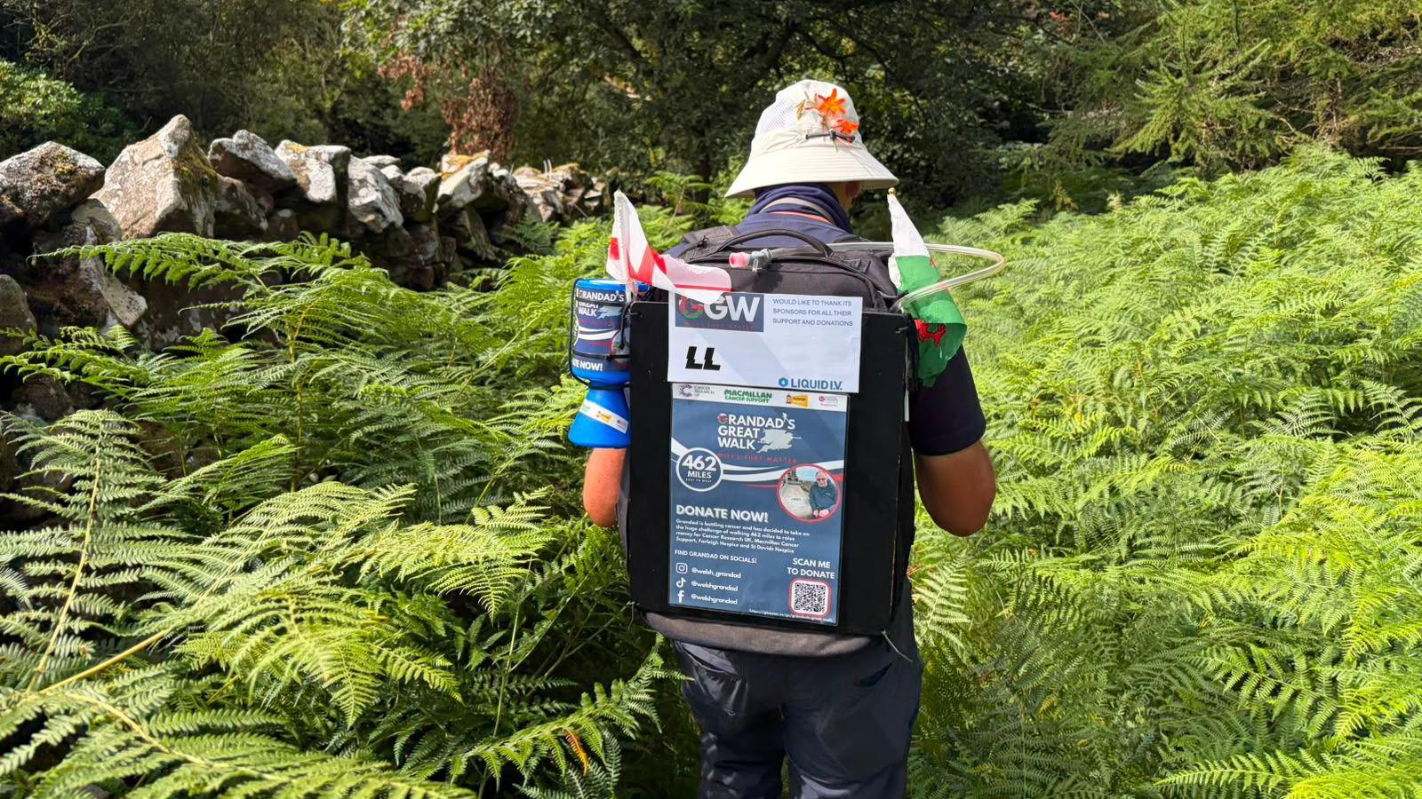 John walks away from the camera through an overgrown footpath full of ferns. He is wearing a backpack that is decorated with a Welsh flag and English flag, as well as the information on how to donate to his fundraising walk.