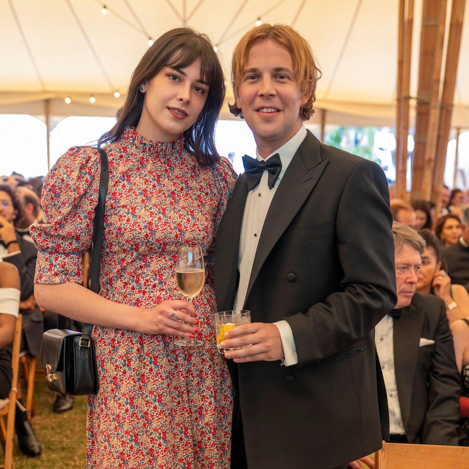 Tom Odell and Georgie Somerville pose at a garden party in 2022, he is wearing a black suit and she is wearing a pink floral dress