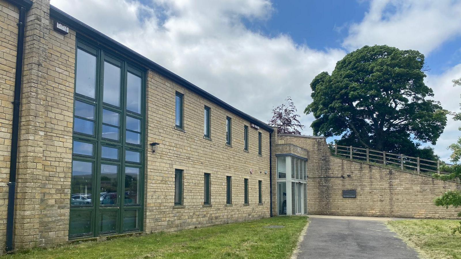 A view of the exterior of The Yorkshire Dales National Park Authority building, which is a large sand-coloured stone building, with green-framed windows and a flat roof.