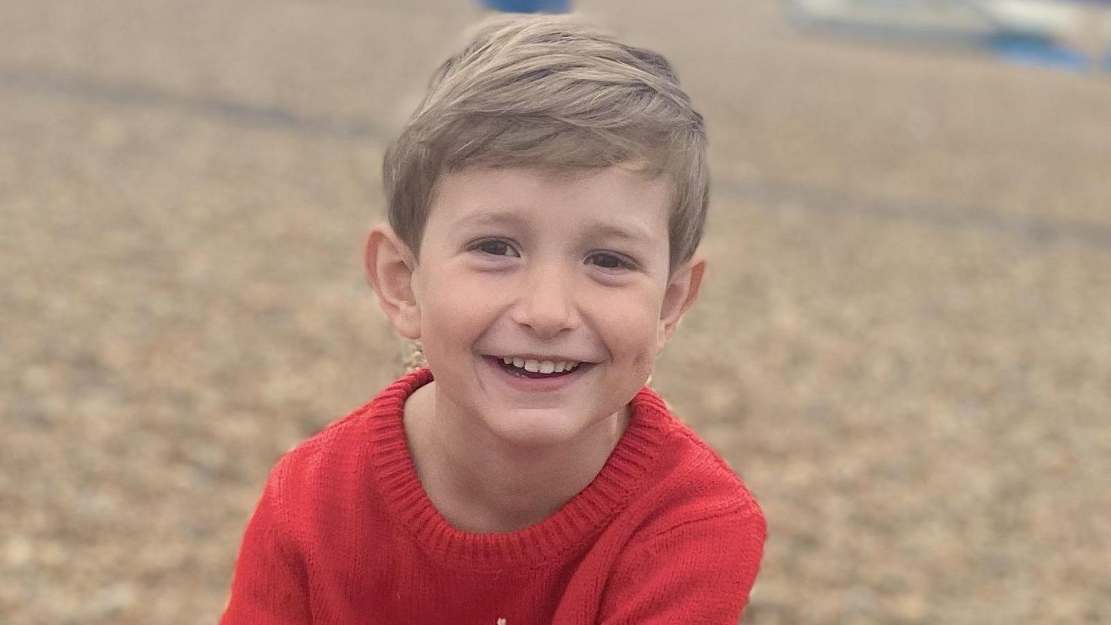 Benedict, a young boy with blond hair, is on a pebbly beach. He is wearing a red knitted top and is smiling to the camera.
