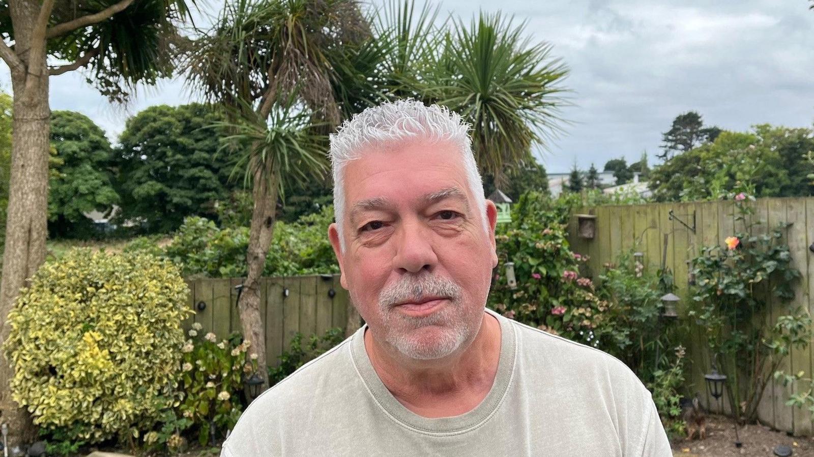 A man stands in his neat garden with flowers, he wears a grey T shirt.