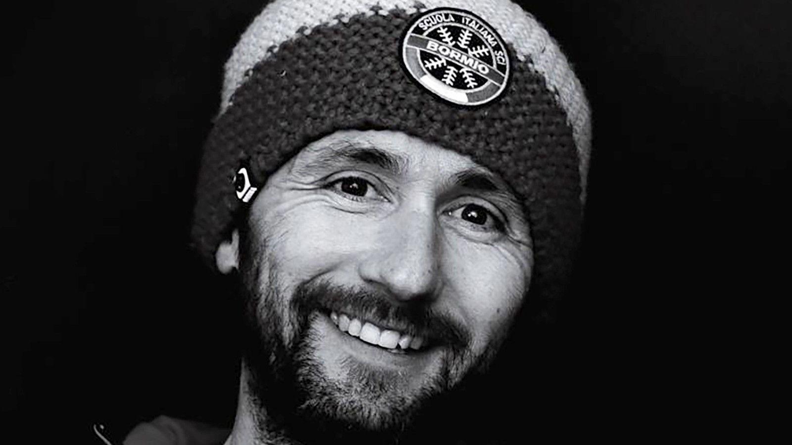 A black and white photo of Justin Loveland. He is smiling broadly, has a beard and is wearing a beanie with Italian words on a badge at the front.