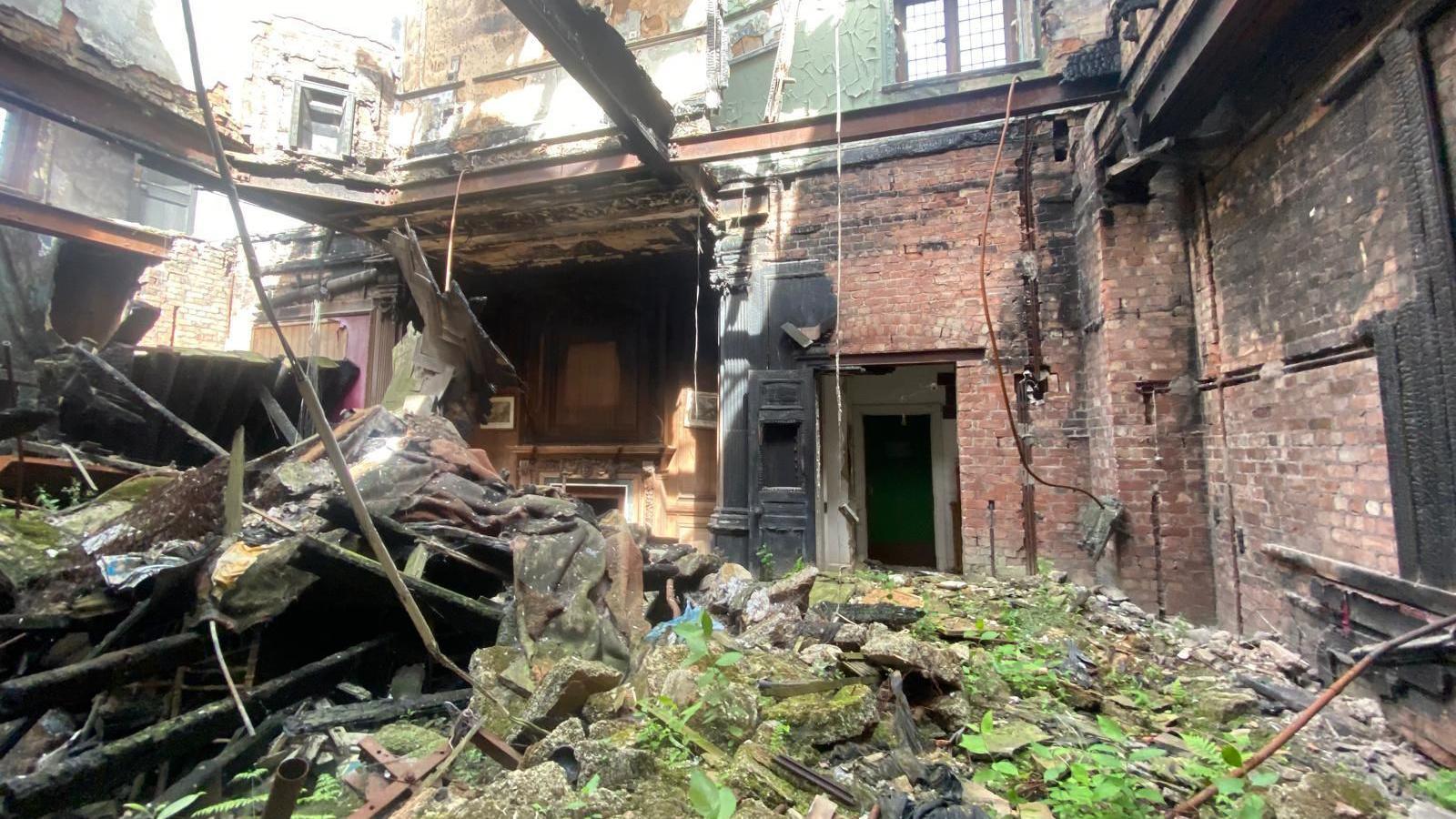A room inside an old manor house. The ceiling has collapsed in, the floor is covered in debris and plants are sprouting out of the ground.