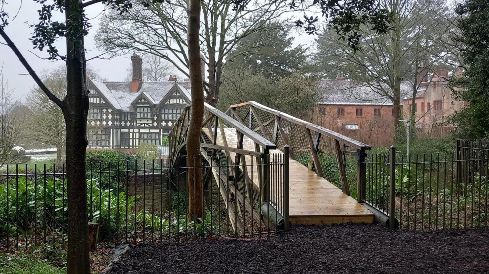 The restored bridge with railings on either side of it and buildings in the background. The wooden bridge is arched and has a new shiny base. 