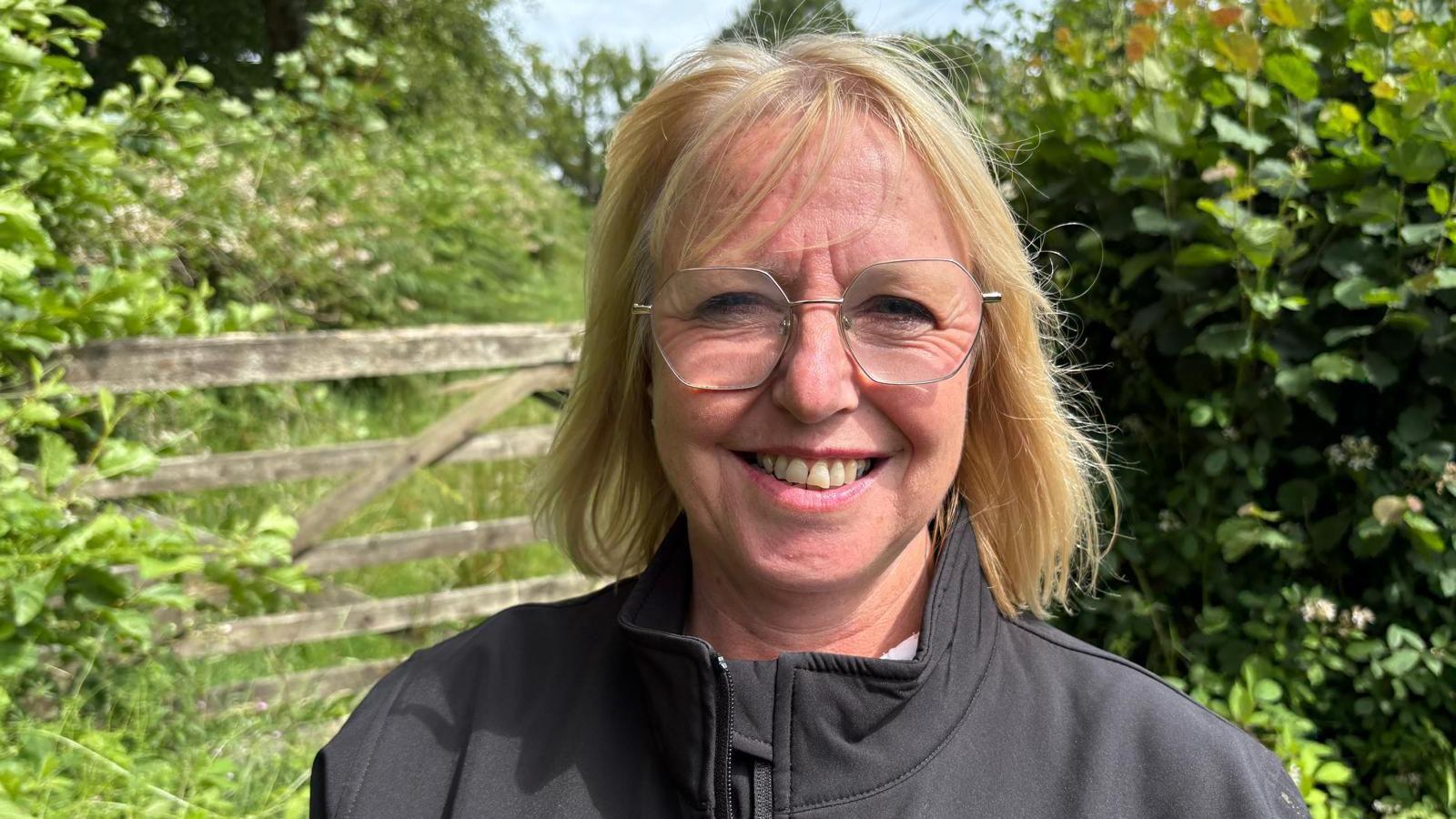Andrea is outside on a sunny day. She is wearing a black, zipped jacket and has shoulder-length blonde hair and glasses. She is smiling at the camera. Behind her is a wooden farm-gate and hedges.