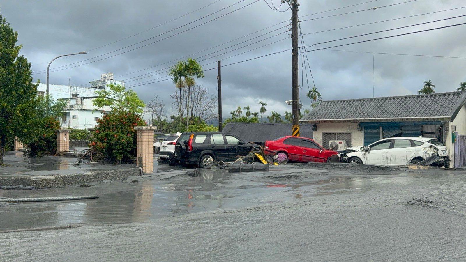 Two damaged cars (one white and one red) in front of a house after floating in the grey floodwater
