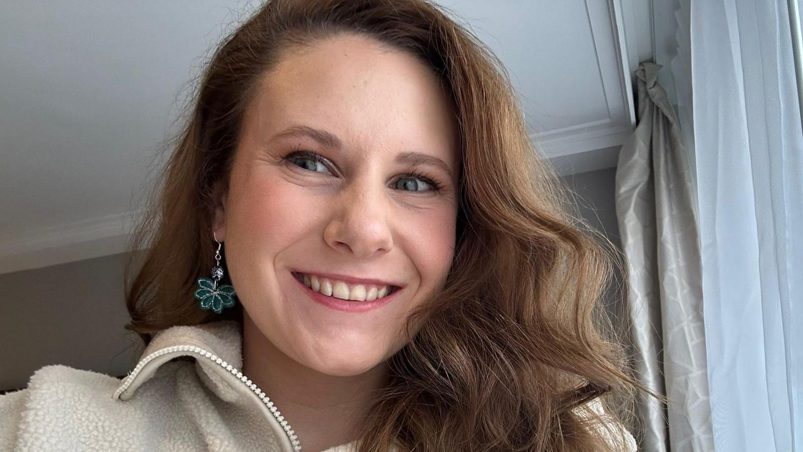 A selfie of Molly Leonard. She has long, brown wavy hair and drop earrings with a turquoise flower on. She is wearing a white fleece. 