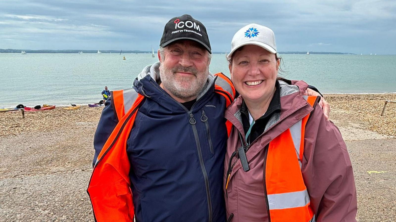 Tony Bray and Catherine McGaw-Pratt standing on a beach and smiling at the camera. They are wearing baseball hats and orange hi-vis vests over their coats