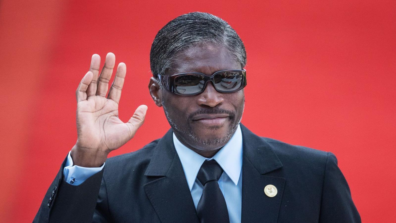 Teodoro Obiang Mangue wearing a dark suit and sunglasses, waving in front of a red background. 