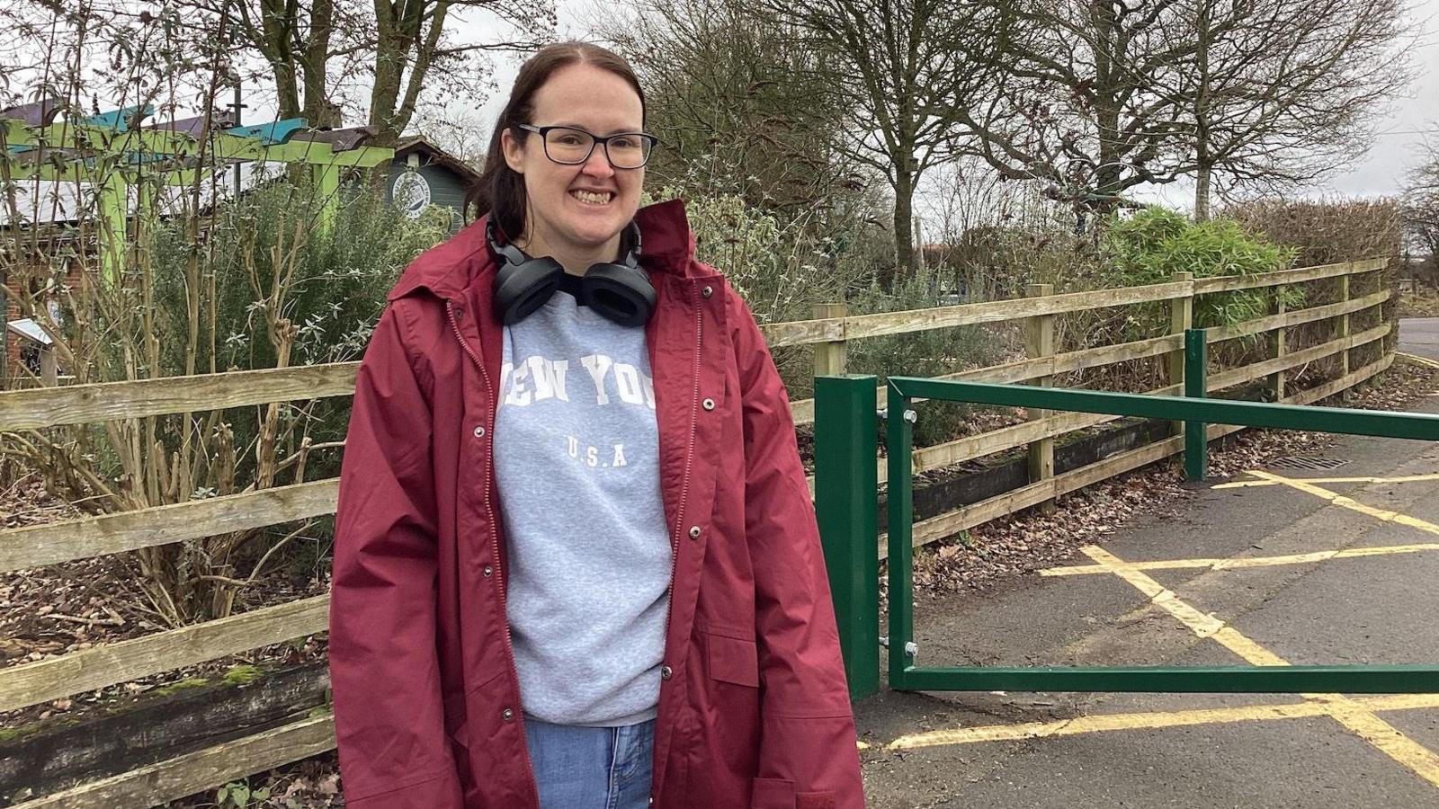 A woman in a red waterproof jacket, grey jumper and blue jeans standing outside of a country park. She has black headphones, hanging around her neck, her hair is tied back and she has black framed glasses on