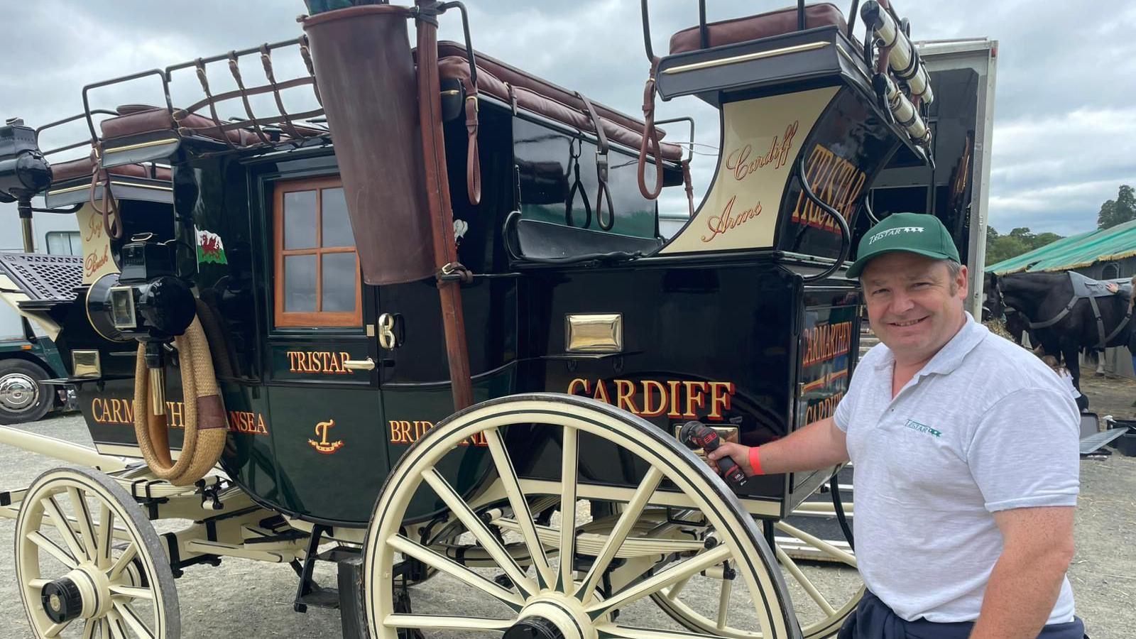 A man pictured next to an old fashioned dark green carriage. The carriage is very shiny and has large cream wheels. The man is standing to the right of the vehicle and is wearing a grey t-shirt and green cap. 