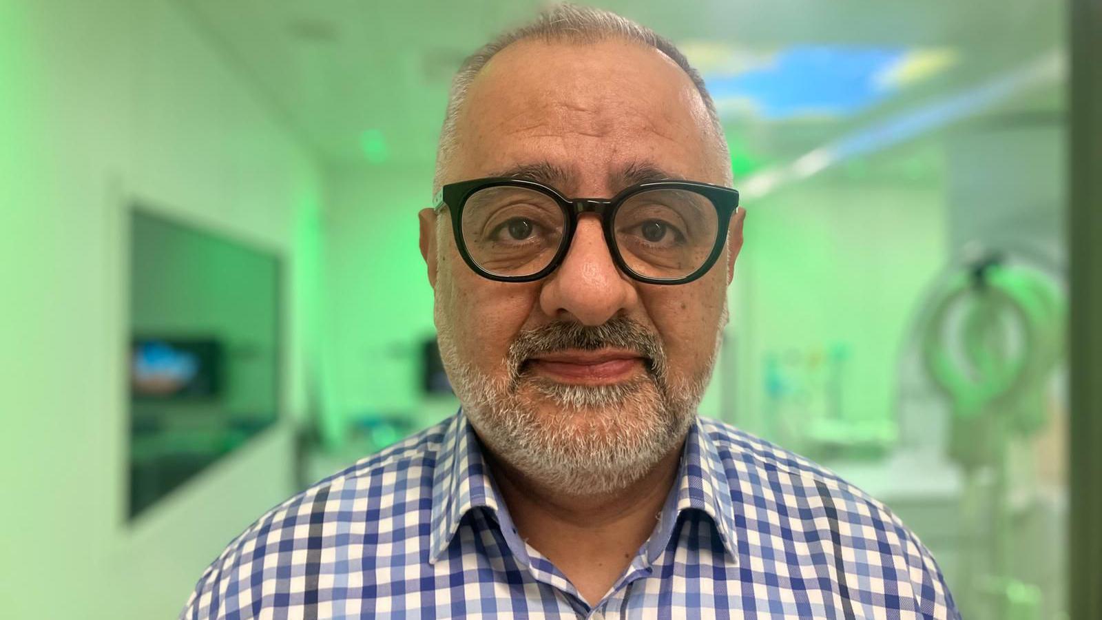 Dr Bashar Al-Qaisieh wears a blue and white gingham shirt with glasses. The MRI scanner is blurred in the background - green lighting illuminates the room it is in.