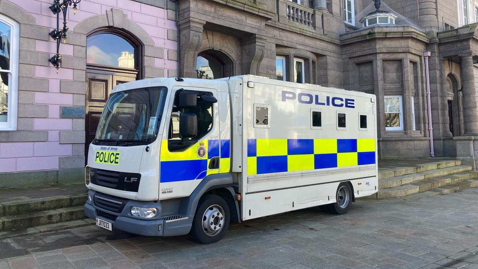 A white States of Jersey Police prison van with blue and yellow rectangles along the side and with police in large navy blue block capitals written near the top of the vehicle. The van is parked in front of buildings with grey and pink bricks.