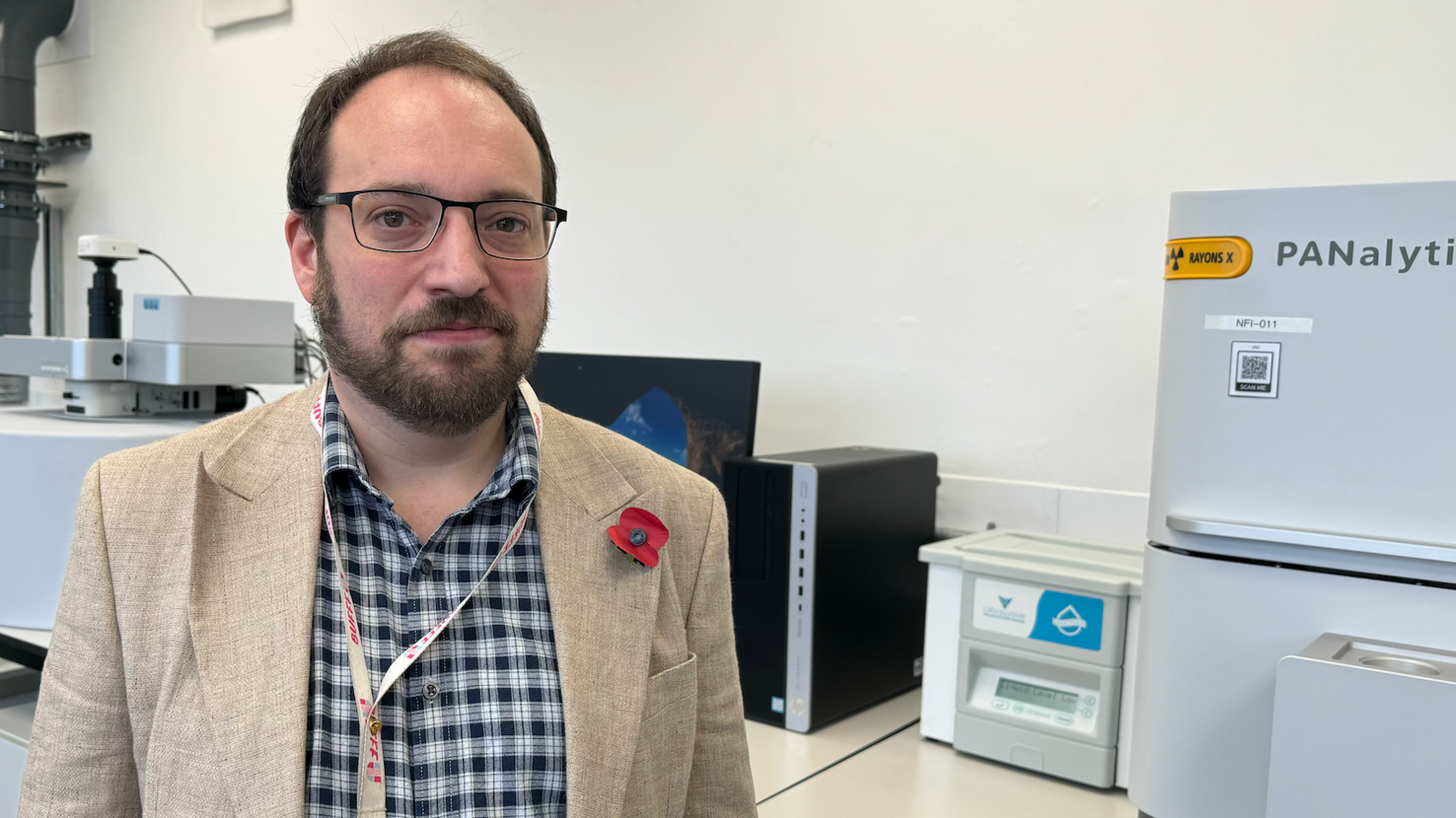 Simon Middleburgh portrait, wearing a checked black and white shirt with a light coloured jacket, wearing a remembrance poppy - situated in a laboratory with scientific machinery behind him