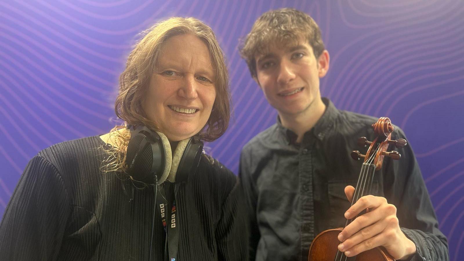 BBC Radio Sussex presenter Sarah Gorrell pictured with Matthew English at the BBC studios in Brighton. Matthew is wearing a navy shirt and is holding his violin in his left hand. Sarah is wearing a black jacket and her headphones can be seen in the image.