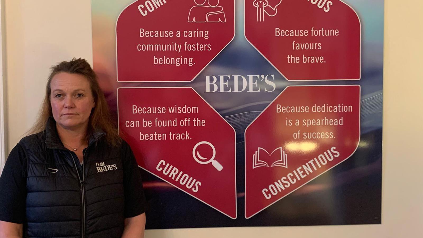 Dani has long brown hair. She stands in front of the St Bede's School logo showing the words compassionate, courageous, curious and conscientious