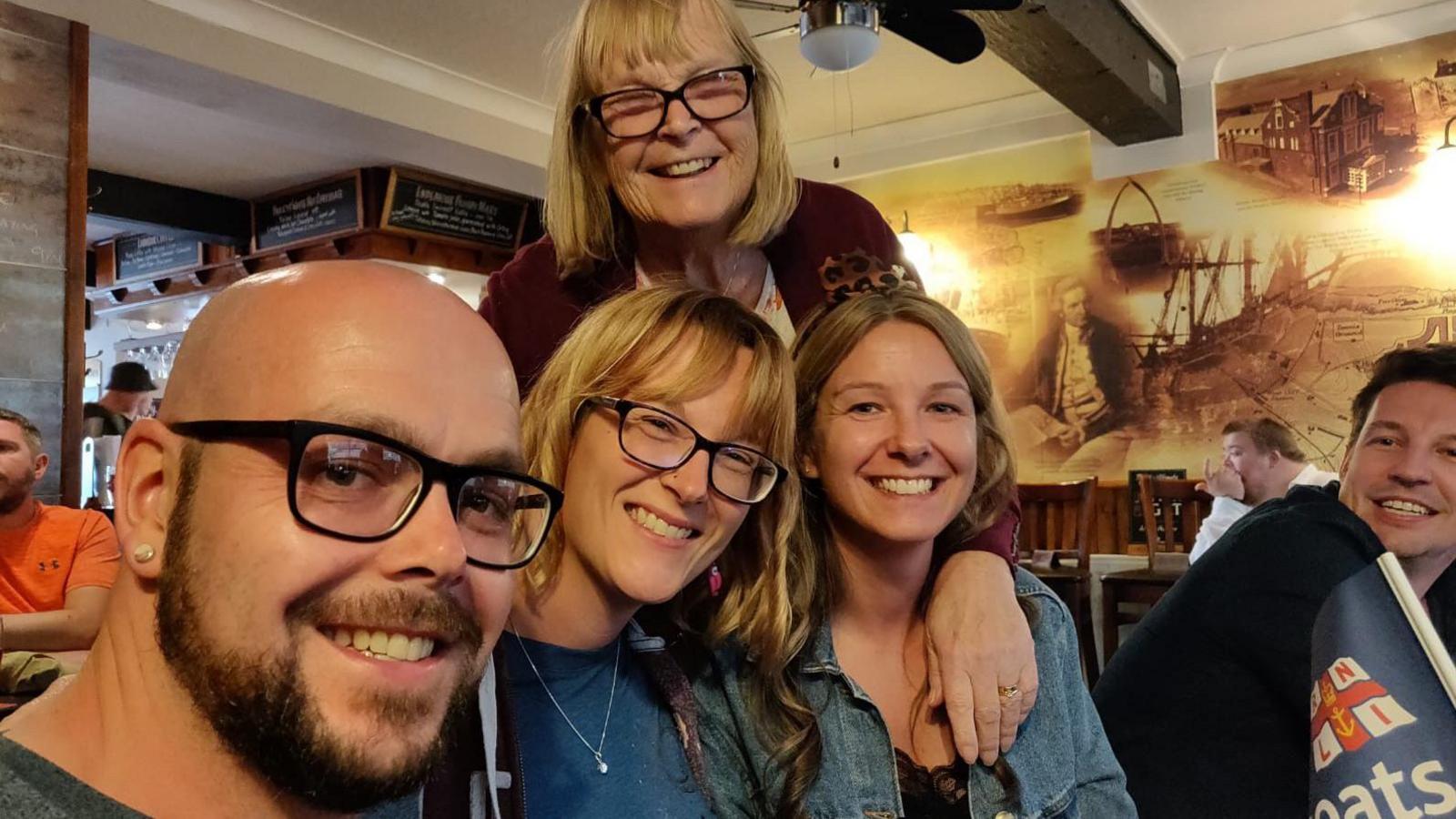 Family group picture of Linda Imrie smiling and standing behind her three adult children, who are sitting down in a restaurant.